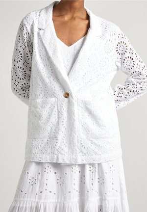 Kvinde iført en hvid broderet blondeblazer med en enkelt knap og matchende hvid broderet blondeskørt.