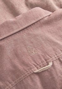 Camicia di tessuto a righe rosa con un piccolo logo ricamato di "&". Include un colletto e un dettaglio con bottoni sul pattino. Texture morbida.