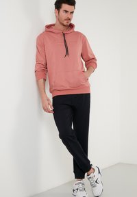 Rózsaszín kapucnis pulóver elülső zsebbel, állítható zsinórral és domborított felirattal, fekete joggingnadrággal párosítva. A modellen fehér és szürke sportcipők vannak.
