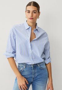 Camisa de botones a rayas azul y blanca, con un corte relajado, mangas enrolladas y un collar clásico, hecha de tela liviana.