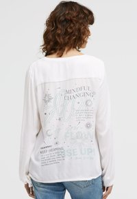 Frau mit lockigem braunem Haar, die ein weißes Langarmshirt mit inspirierenden Texten und grafischen Designs auf dem Rücken trägt, kombiniert mit blauen Jeans.
