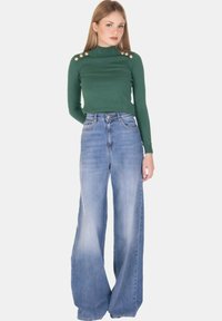 Maglione verde a coste aderente con dettagli in bottone sulle spalle, abbinato a jeans in denim azzurro chiaro a gamba larga. Texture liscia e design casual.