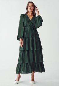 Green, tiered maxi dress made from flowing fabric. Features a V-neck, long puff sleeves, and a tie waist. Accented with ruffles.