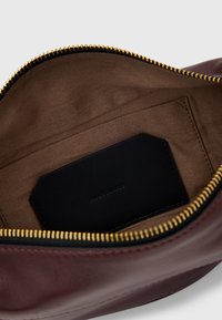 Interno di borsa in pelle bordeaux con una texture liscia, zip dorata e fodera in tessuto marrone; include un'etichetta in pelle nera.