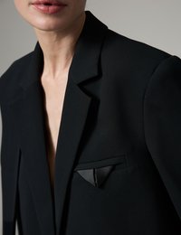 Κοντινό πλάνο ατόμου που φοράει μαύρο σακάκι με μαντηλάκι τσέπης με σατέν λεπτομέρεια, δείχνοντας τον λαιμό και τα χείλη, σε ουδέτερο φόντο.