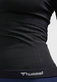Schwarzes, langärmliges Sportshirt mit gerippter Textur und Hummel-Logo am Saum. Figurbetontes Design mit dezenten Musterdetails.