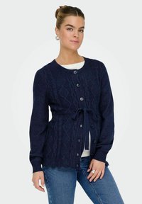 Tamno plavi pleteni kardigan s teksturiranim dijamantnim uzorkom, pet dugmadi sprijeda i detaljem vezivanja u struku. Dugi rukavi s rebrastim manžetama.