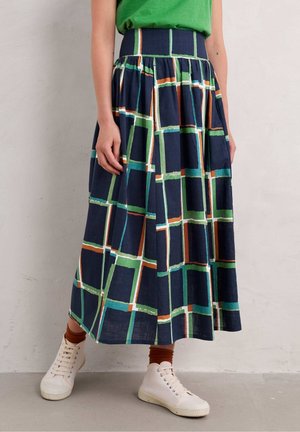 Femme portant une jupe taille haute bleu marine avec un motif à carreaux vert, blanc et orange, associée à des chaussettes marron, des baskets blanches et un haut vert.