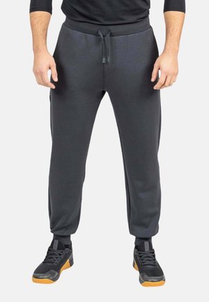 Hombre con pantalones deportivos gris oscuro con cordón y zapatillas negras con suelas naranjas, de pie frente a un fondo blanco.