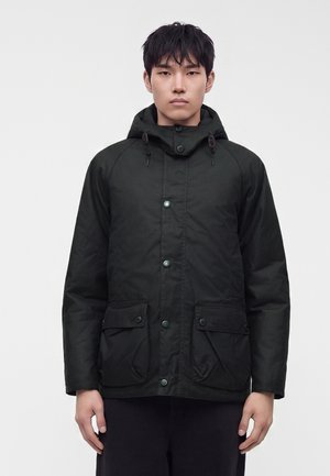 Young man wearing a black hooded jacket with buttoned front and flap pockets, standing against a plain light background.