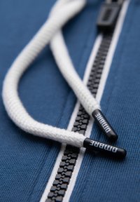 Gros plan sur une fermeture éclair noire sur un tissu bleu avec des cordons blancs tressés, chacun se terminant par des embouts en plastique noirs portant la mention "DIMBRE".