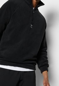 Schwarzer Fleecepullover mit einem Reißverschluss in Viertellänge, gerippten Bündchen und einem kleinen weißen Logo auf der Brust. Weiche Textur und lässige Passform.