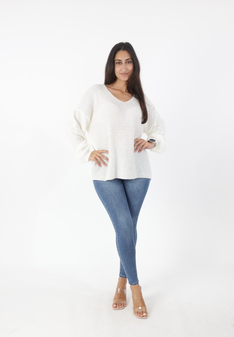 Witte gebreide trui met een V-hals en brede mouwen, gecombineerd met strakke blauwe jeans en doorzichtige sandalen met hakken. Effen achtergrond.