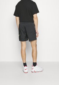 Man med svarta shorts, svarta strumpor, vita och röda Nike-sneakers, och en svart t-shirt, står vänd mot en vit vägg.