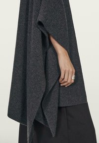 Ruka částečně viditelná skrze rozparek ve tmavě šedém pleteném ponču, které je nošeno přes černou sukni, se stříbrným prstenem na prstu.