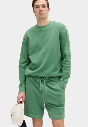 Homme portant un sweat-shirt vert à manches longues et un short vert assorti, tenant une casquette de baseball blanche dans la main gauche.
