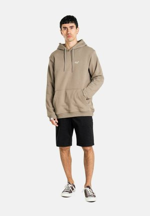 Junger Mann trägt einen taupefarbenen Hoodie mit dem Schriftzug "Loyal", schwarze Shorts und schwarz-weiße Sneakers, steht mit einer Hand in der Tasche vor weißem Hintergrund.