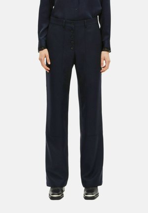 Vrouw in een marineblauwe wijde broek met knopen aan de voorkant, gecombineerd met zwarte schoenen en een marineblauw blouse met kant afgewerkte mouwen.