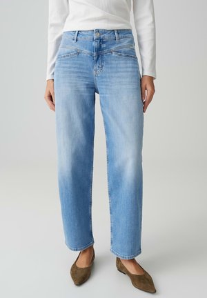 Femme portant un jean droit bleu clair, un haut blanc à manches longues côtelé, et des ballerines pointues en daim marron, debout devant un fond uni.