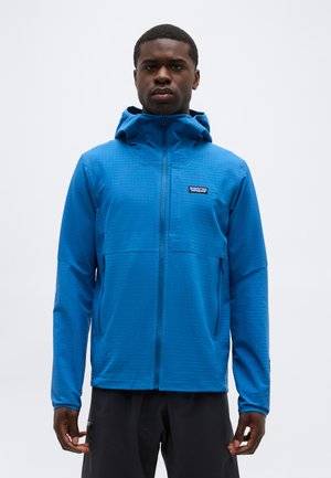 Homme debout, face à l'avant, portant une veste à capuche zippée bleue avec le logo Patagonia et un pantalon noir, sur un fond uni.