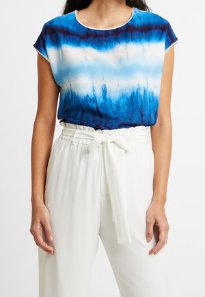 Kobieta w niebiesko-białej bluzce tie-dye, włożonej do białych spodni z wysokim stanem, z materiałowym paskiem związanym kokardą.