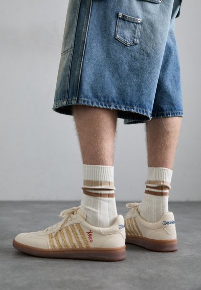 Sneaker retro beige con suola in gomma marrone, dotate di dettagli a righe e loghi ricamati. Indossate con calze bianche e dettagli marroni a righe.