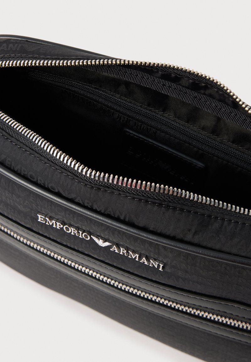 Schwarze Reißverschlusstasche mit geprägt außenliegendem Logo, glatter Textur und metallischen Beschlägen. Innen verfügt sie über ein Stofffutter und zusätzliche Taschen.