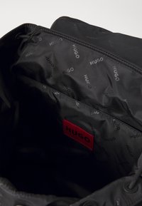 Black fabric bag interior with repeated "HUGO" logo pattern. Features a red label with "HUGO" text, drawstring closure, and smooth texture.