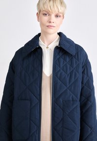Navy blue quilted jacket with diamond pattern, large collar, two front pockets, and silver zipper, layered over a cream shirt and beige vest.