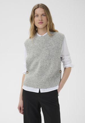 Femme aux cheveux blonds portant un pull sans manches gris en tricot par-dessus une chemise blanche et un pantalon noir, debout avec une main dans la poche.