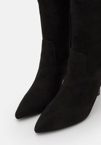 Bottes hautes jusqu'au genou en daim noir avec un bout pointu et une fermeture éclair latérale. Elles présentent un design élégant et un petit talon empilé.