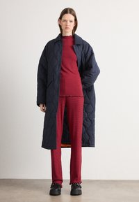 Cappotto lungo trapuntato blu navy sopra una maglietta rossa a coste e pantaloni abbinati. Scarpe nere chunky. Colori solidi senza motivi o accenti visibili.