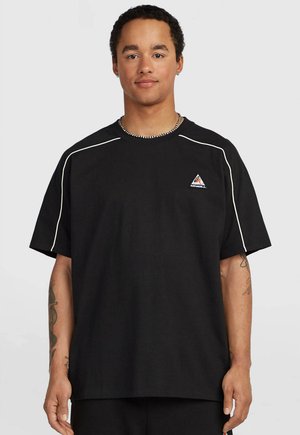 Young man with short curly hair wearing a black oversized t-shirt with white piping and a small triangle logo on the chest.