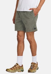 Mannelijke groene shorts van zacht, gestructureerd stof met zijzakken; heeft een zwart rond logo. Gecombineerd met bruine sportschoenen en witte sokken.