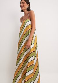 Robe maxi avec des rayures diagonales vertes, jaunes et rouges sur fond blanc. Fabriquée en tissu léger avec de fines bretelles spaghetti.