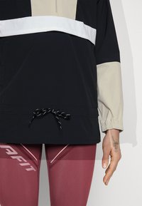 Μαύρος και μπεζ πουλόβερ με μισό φερμουάρ, ελαστικούς μανσέτες και ρυθμιζόμενα κορδόνια. Φορεμένο πάνω από μπορντώ κολάν με εκτυπωμένα στοιχεία.