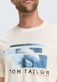 Hombre con camiseta beige que tiene un gráfico en azul y blanco y el texto "Tom Tailor High Peak Avalanche" en el frente.