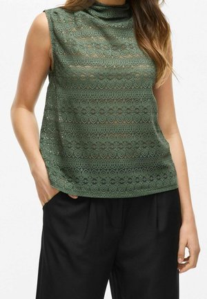 Haut sans manches en dentelle vert olive à col montant, associé à un pantalon noir ample, main du mannequin dans la poche, fond neutre.