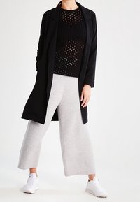 Svart kappa med öppen front över en svart mesh-top, kombinerad med ljusgrå vida byxor och vita sportskor. Släta, andningsbara texturer.