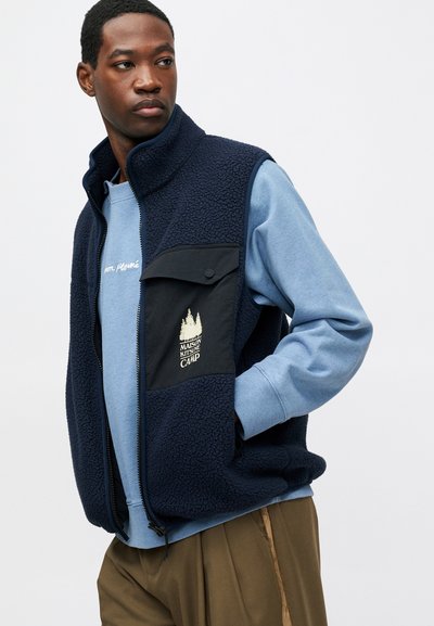 Námornícky fleece vest s vysokým golierom, čiernou náprsnou kapsou a zlatým logom. Nosený cez svetlomodrú mikinu, v kombinácii s hnedými nohavicami.