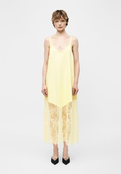 Jeune personne portant une robe jaune pâle sans manches avec un ourlet en dentelle et des chaussures noires à bout pointu, debout devant un fond clair uni.
