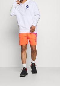 Vit hoodie med blå Nike-logotyp, långärmad med gul-gröna accenter. Klara orange shorts med grafisk tryck. Svarta sneakers.