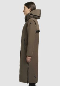 Cappotto puffer lungo marrone con colletto alto e cappuccio. Presenta tasche con zip e un logo triangolare sull'orlo. Texture liscia, design elegante.