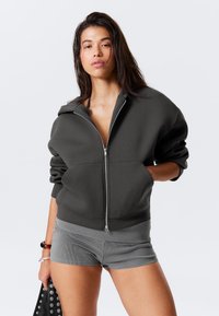 Dark gray zip-up hoodie with a relaxed fit, large front pockets, and a soft texture. Paired with gray ribbed shorts.