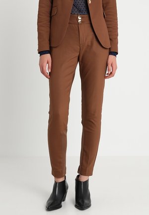 Frau trägt braune maßgeschneiderte Hose mit zwei Knöpfen, passenden braunen Blazer, dunkelblaues gepunktetes Hemd und schwarze Stiefeletten.