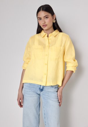 Gul linned skjorte med knap-up foran, to brystlommer og opkrøllede ærmer, parret med lyseblå jeans.