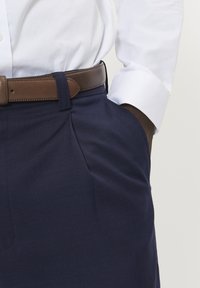 Granatowe spodnie o gładkiej fakturze, z przeszytymi zakładkami i brązowym skórzanym paskiem, noszone z białą koszulą zapinaną na guziki.