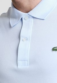 Helles blaues Poloshirt aus strukturiertem Baumwollstoff, mit einer Knopfleiste mit drei Knöpfen und einem kleinen grünen gestickten Logo.
