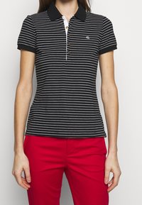 Pruhované černo-bílé polo tričko s krátkým rukávem, límečkem a třemi knoflíky, s logem na levé hrudi. Viditelné červené kalhoty.