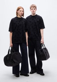 Μαύρα υπερμεγέθη T-shirts με υφασματικές υφές, συνδυασμένα με σκούρα τζιν παντελόνια καμπάνα. Τα μοντέλα κρατούν μαύρες τσάντες με διάφορα σχέδια.